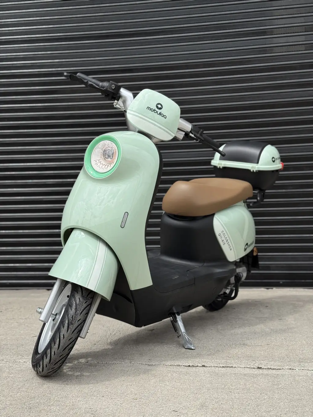Scooter eléctrico estilo retro color verde menta y negro con batería de litio, estacionado frente a persiana metálica.