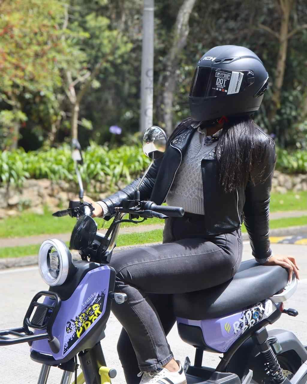 Motociclista con casco DOT negro y chaqueta de cuero sobre scooter eléctrico.