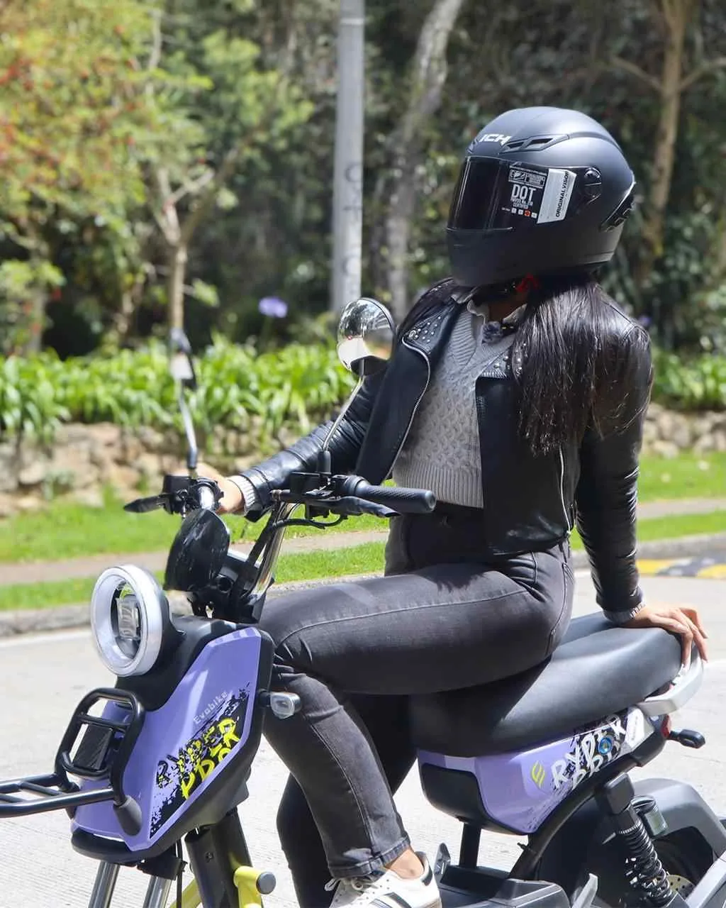 Mujer con casco negro y chaqueta de cuero disfrutando su eBike morada en la ciudad, ejemplo de movilidad eléctrica y seguridad vial.