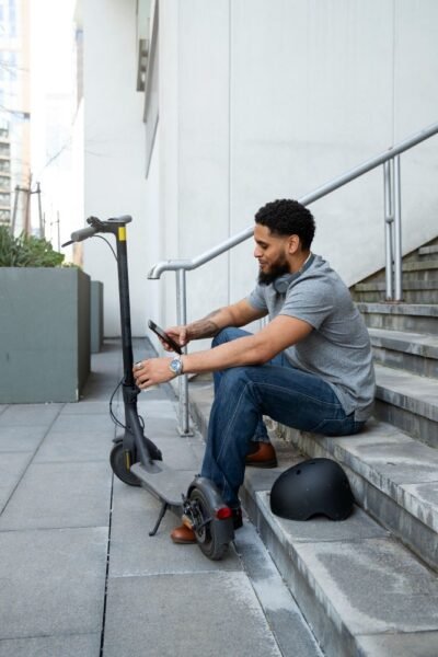 Hombre sentado junto a su ciclomotor eléctrico en entorno urbano, revisando su teléfono antes de continuar el trayecto, símbolo de movilidad moderna y ecológica.